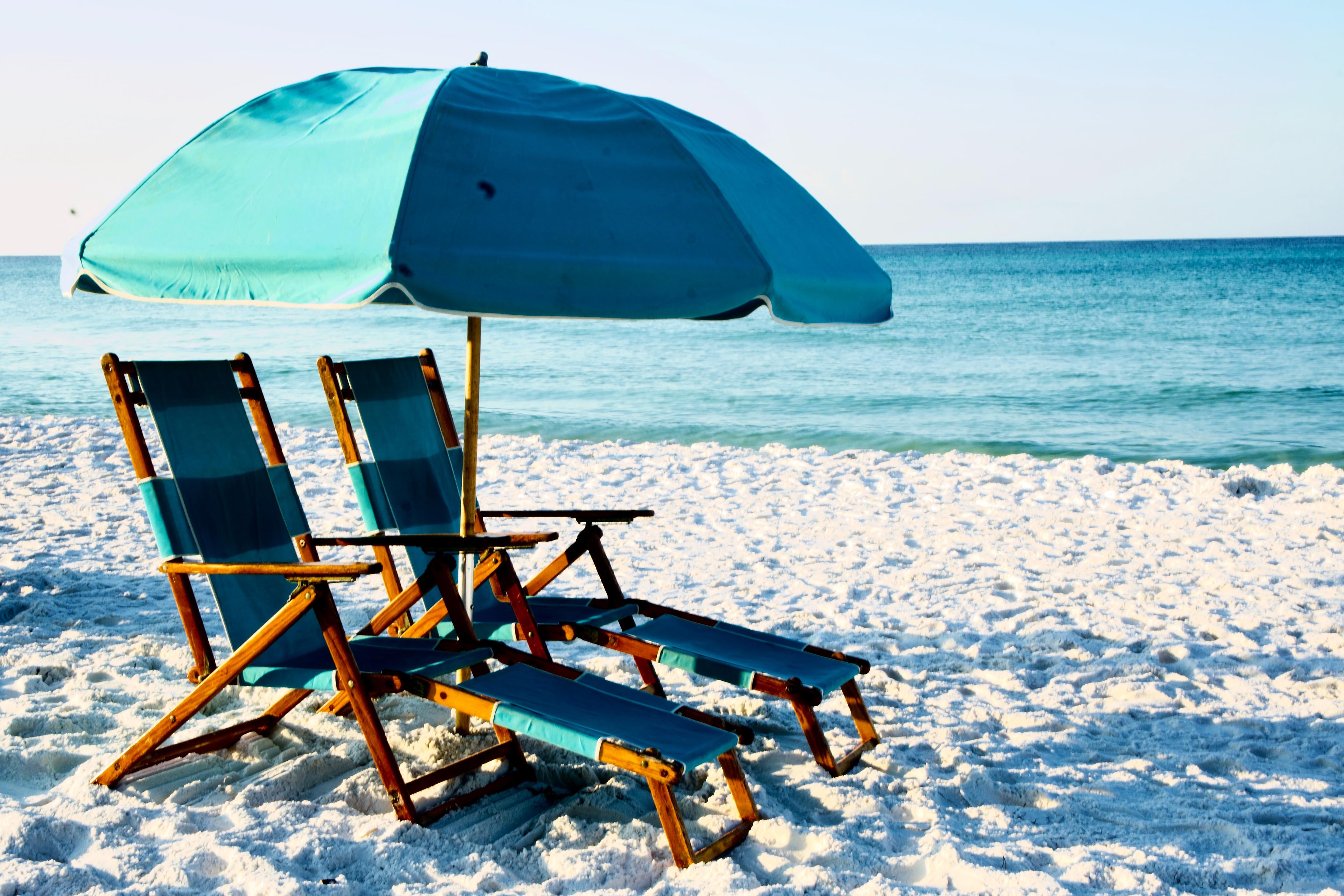 A pair of teal beach chairs under a similarly colored umbrella next to the ocean on a white sandy beach in Destin Florida. 