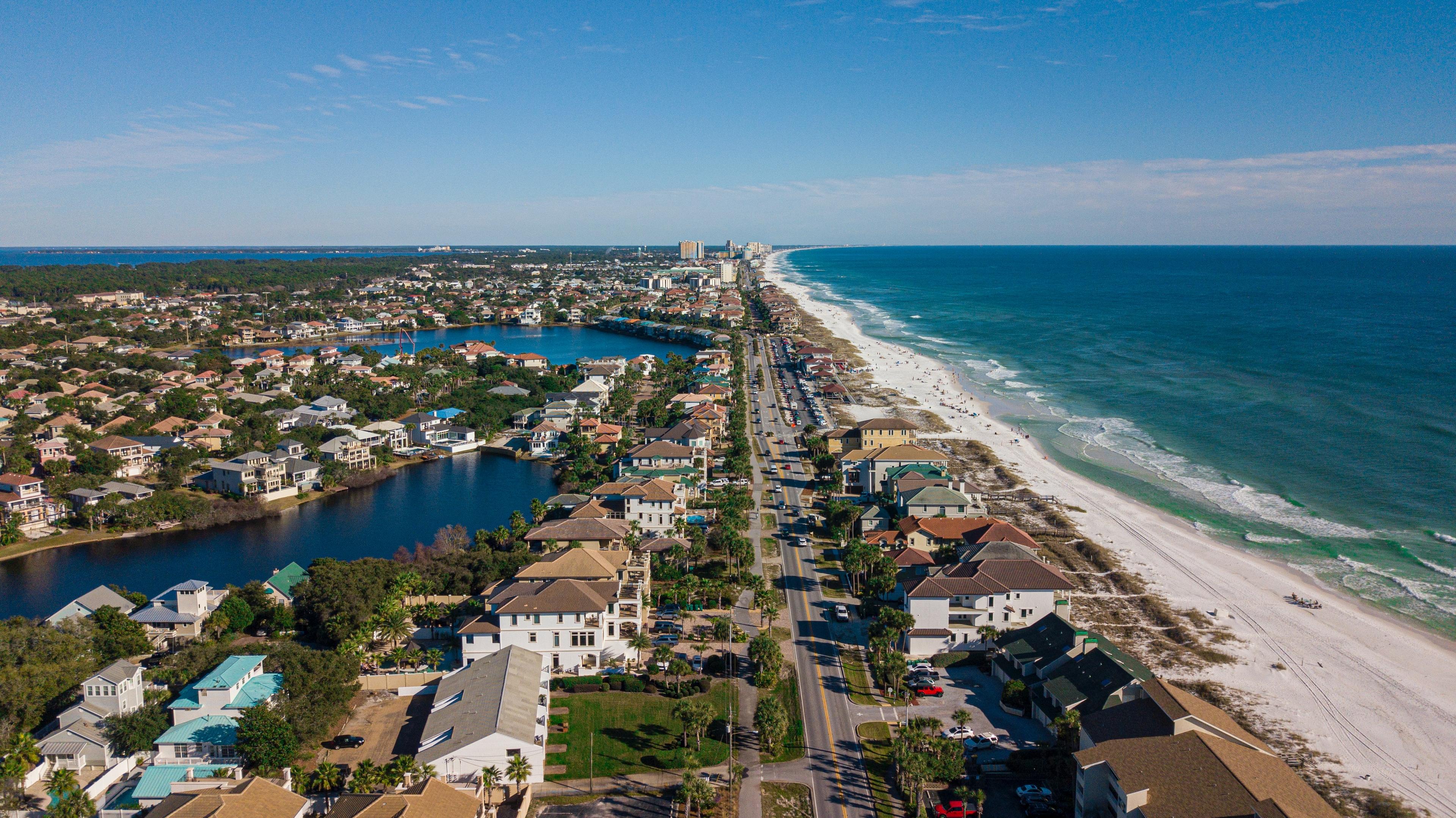 An aerial drone shot of Destin with the road full of cars towards the middle right with houses on either side, to the left is a river and lake and to the right is a beach and the ocean.