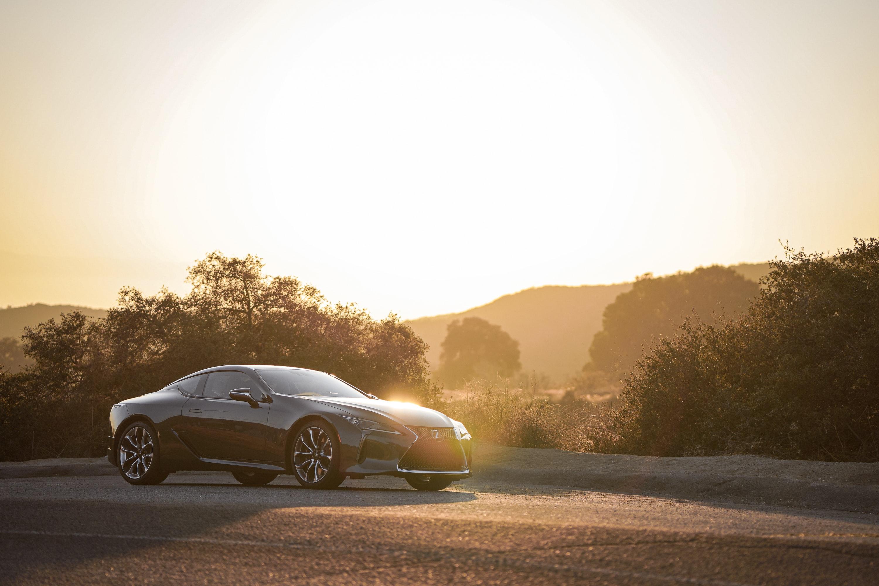 View of a black LC Lexus driving from left to right in front of trees and mountains that are black due to the sun setting and everything is golden.