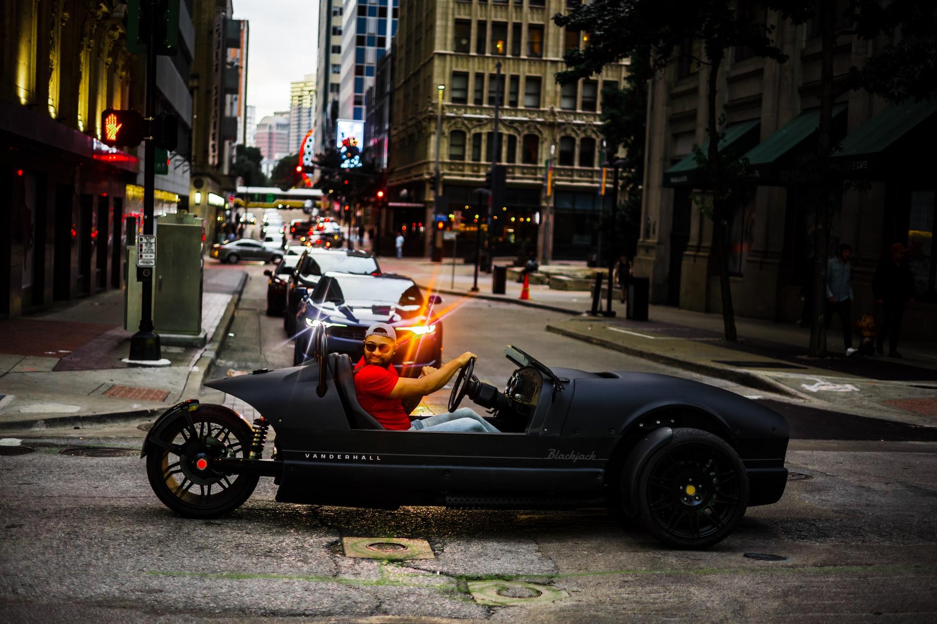 A man driving a black Vanderhall vehicle through city streets