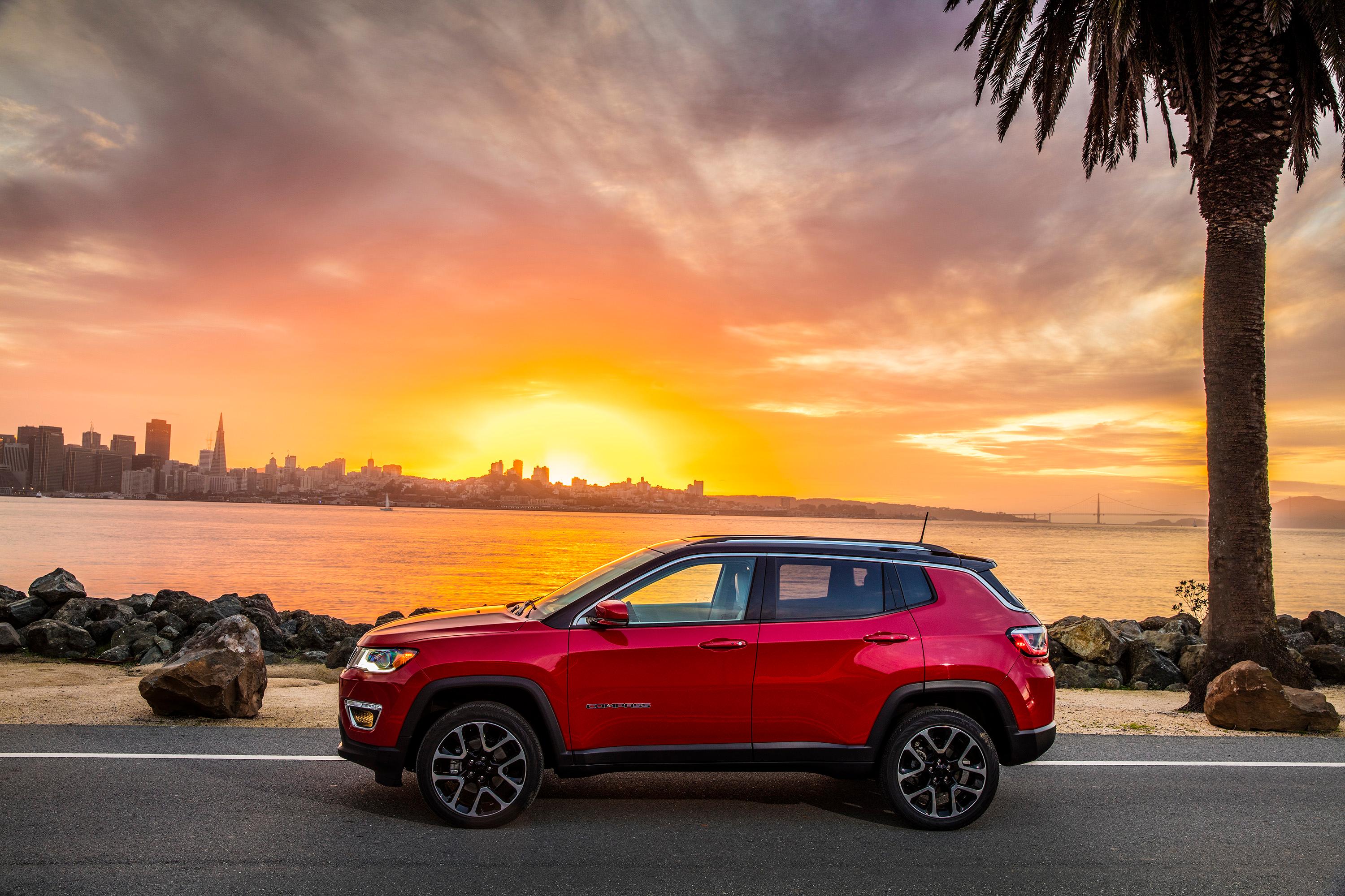Image of a red Jeep Compass driving past a palm tree and large body of water during sunset.