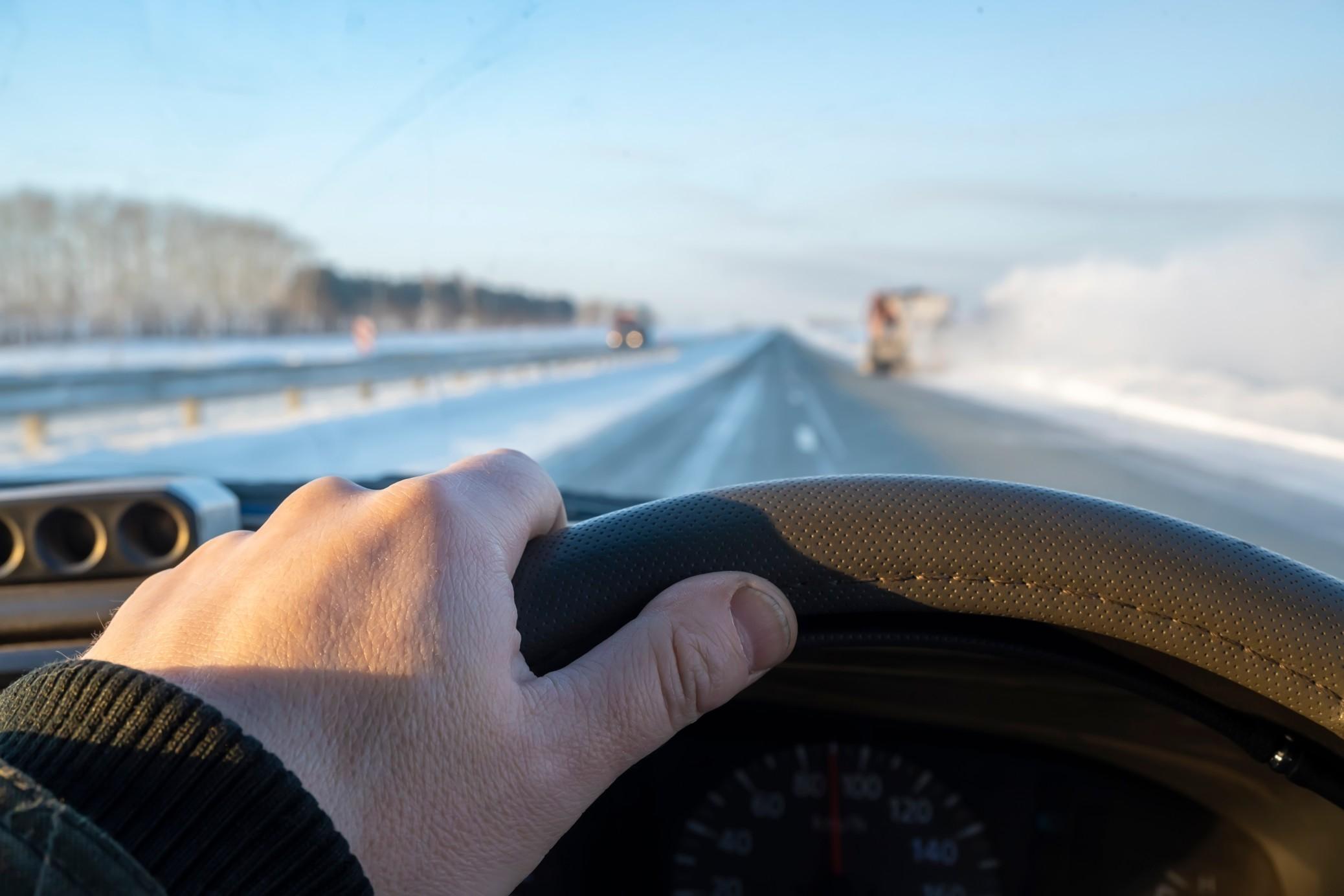 A driver's hand on a steering wheel, driving on a snowy highway.