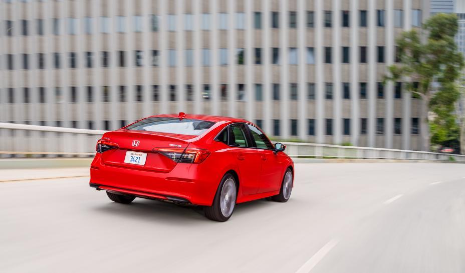 A red 2022 Honda Civic drives along the curve of a highway. In the background, there is a blurry office building and one tree planted alongside the highway.