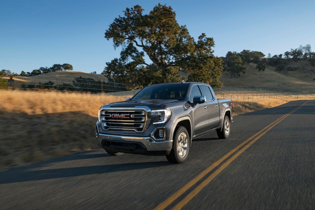 The 2021 GMC Sierra drives down a sunny country road. There is a wheat field to the left of it, and several rolling hills behind it. An olive tree is prominent in the background.