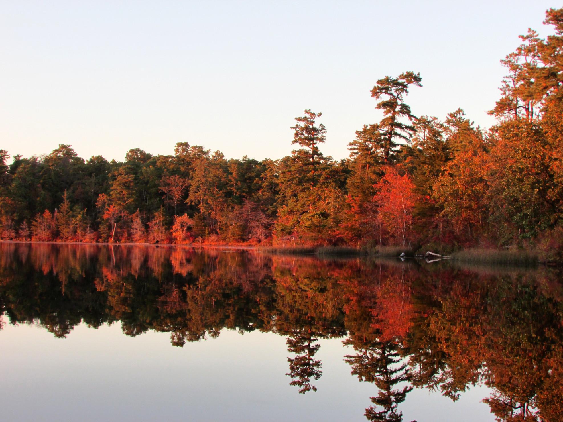 View of a lake surrounded by red and brown trees, reflected in the lake. 