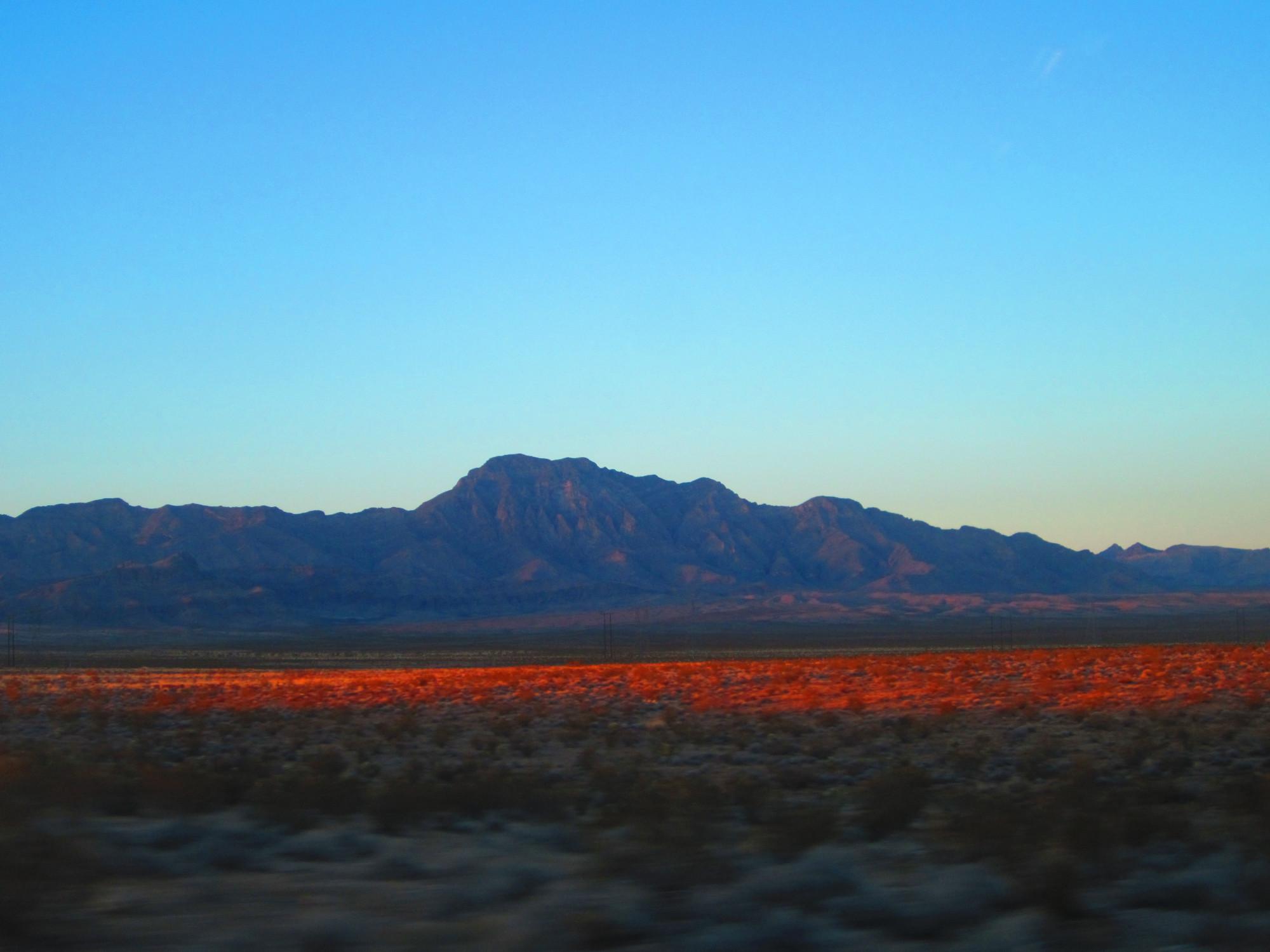 Sunset in the desert, mountain ranges in the background with a vast desert in the front with a strip of red in the middle, presumably the sunset coming through mountains unseen on the right. 