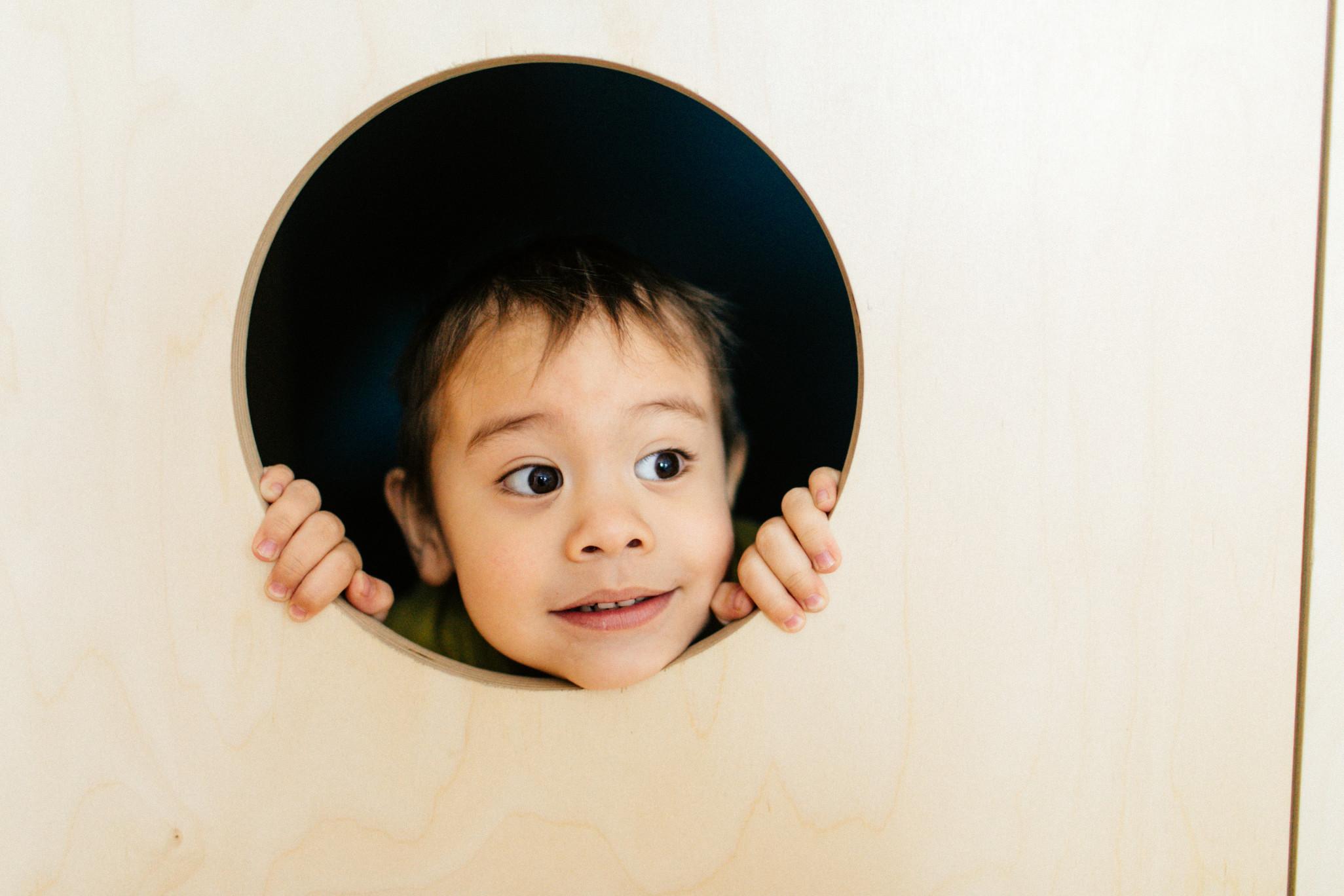 A young boy sticking his head out of a hole in a pale wood play space.
