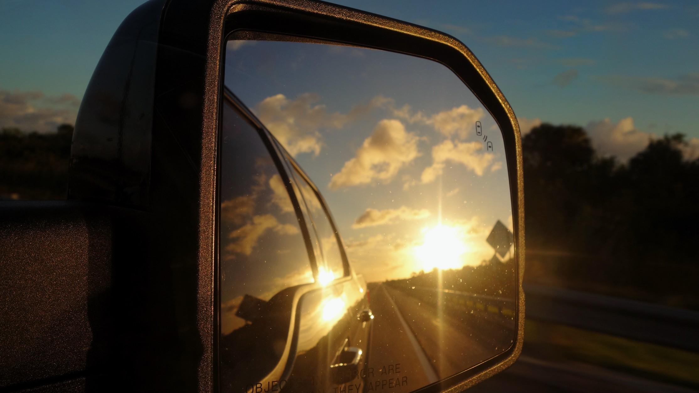 Closeup of a side view mirror with the sun setting in the mirror's reflection. 